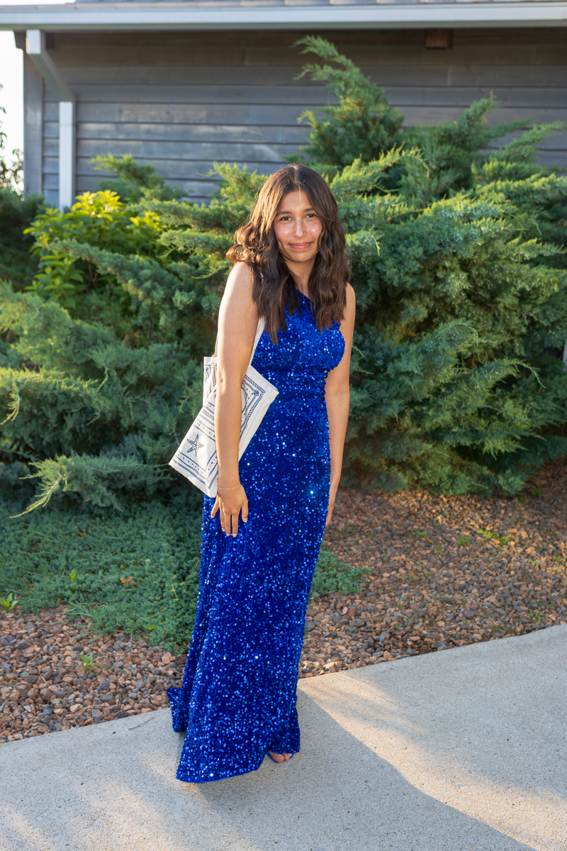 Young woman in a blue sequined evening gown standing on a sidewalk outdoors, smiling with a white tote bag over her shoulder.