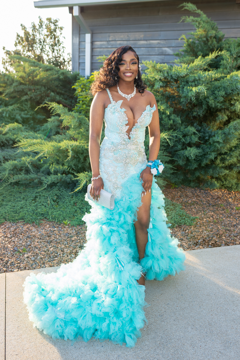 Young woman in a light turquoise, beaded gown with a deep V neckline, posing outdoors among greenery with a silver clutch in hand.