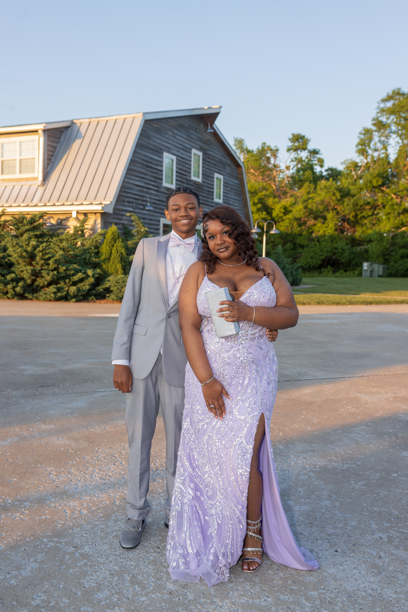 Couple in formal attire posing outdoors: man in light gray suit with bow tie and woman in a lilac beaded gown holding a silver clutch.