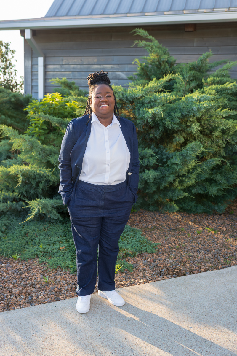 Smiling woman in a navy suit and white shirt stands on a sidewalk with green shrubs and a wooden building behind.