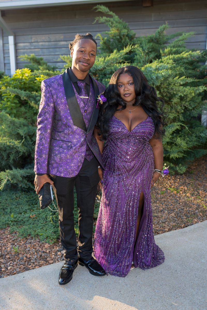 Couple dressed in matching purple formal wear posing outdoors among greenery, smiling at the camera.