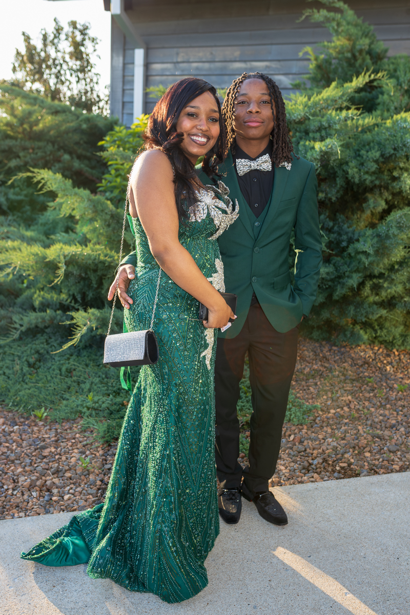 Two people posing outdoors in formal attire: a woman in an emerald beaded gown with silver embroidery and a small silver clutch, beside a man in a green suit and silver bow tie, smiling at the camera.
