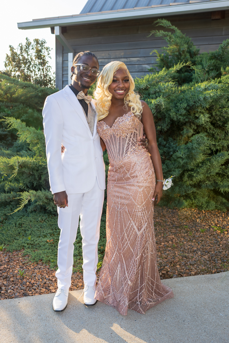 Couple posing outdoors, man in a white suit and bow tie beside a woman in a blush pink sequined gown, both smiling with greenery behind them.