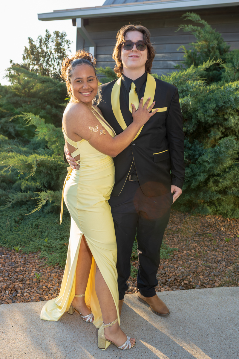 Couple posing outdoors in formal wear: woman in a pale yellow gown embracing a man in a black suit with yellow accents, both smiling.