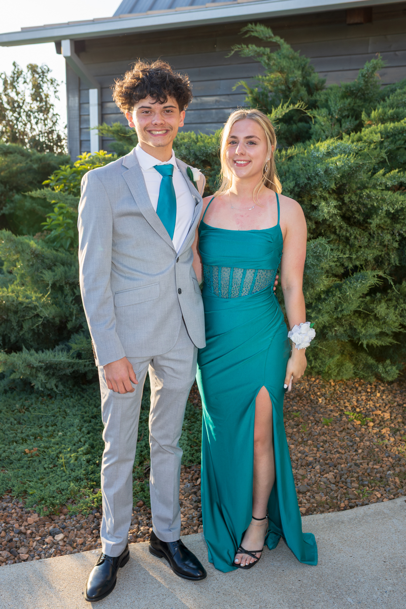 Young couple dressed for a formal event posing outdoors; man in a light gray suit with teal tie and boutonniere, woman in teal gown with corsage wristlet and slit.