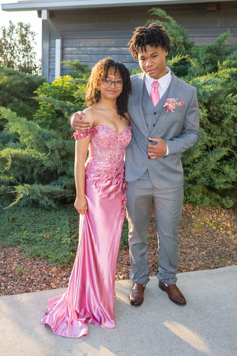 Young couple in formal attire posing outdoors; girl in pink gown, boy in gray suit with pink boutonniere, arms around each other.