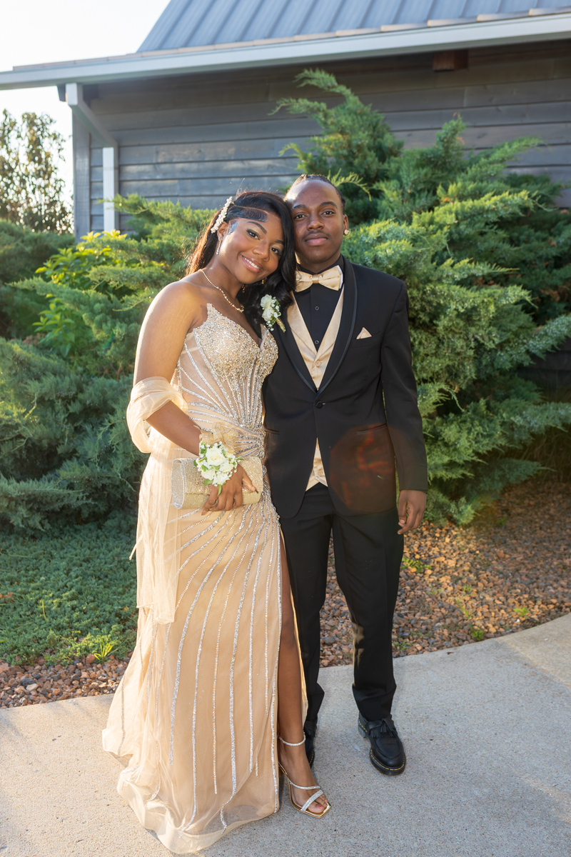 Smiling couple in formal attire posing outdoors; woman in a gold beaded gown with a corsage and clutch, man in a black tux with a gold vest and bow tie in front of shrubs and a building garden backdrop