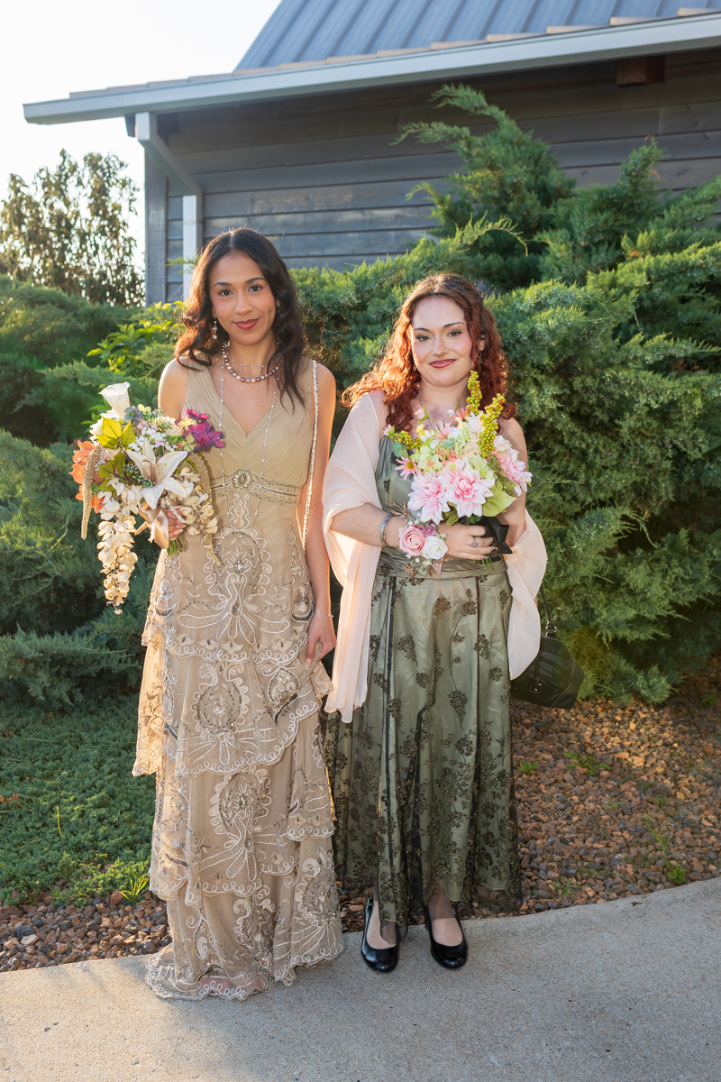 Two women in formal gowns stand on a sidewalk with greenery behind them, each holding a bouquet of flowers.