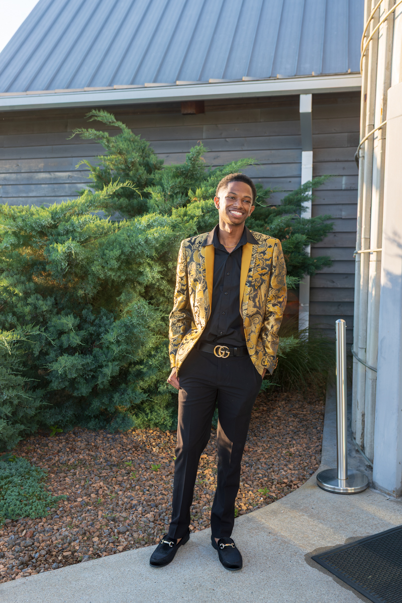 Young man in a gold patterned blazer and black outfit posing outside a building with greenery behind him.