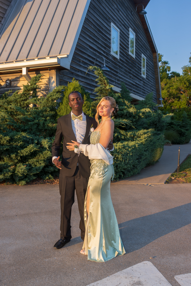 Couple in formal attire posing outdoors beside a wooden building and green shrubs at sunset; man in tuxedo, woman in pale green gown with slit and shawl.