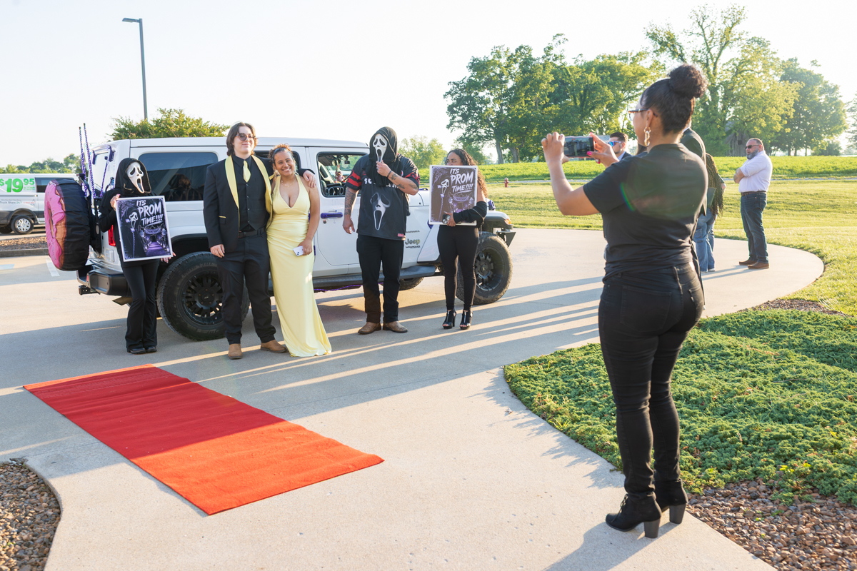 Group posing beside a white SUV on a red carpet; two people wear Ghostface masks and others in formal/costume outfits pose for photos.