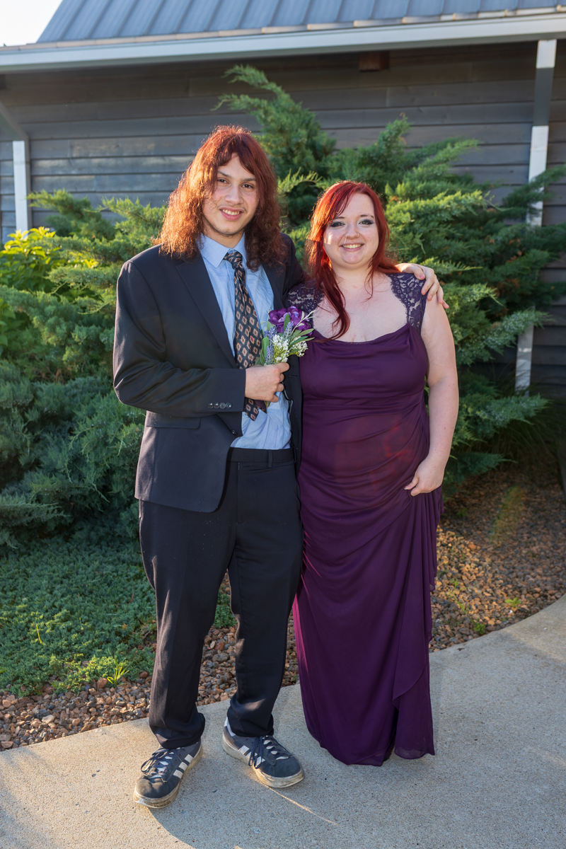Two people posing outdoors: a man in a suit holding a boutonniere and a woman in a purple gown, smiling with their arms around each other in front of greenery.