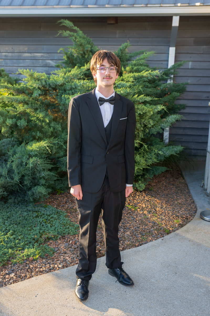 Young man in a black suit with a bow tie standing outdoors near green shrubs and a wooden building backdrop.