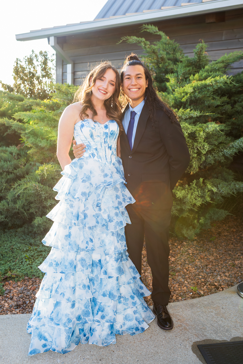 Two friends pose together outside, the woman in a blue floral ball gown and the man in a black suit with a blue tie, smiling at the camera.