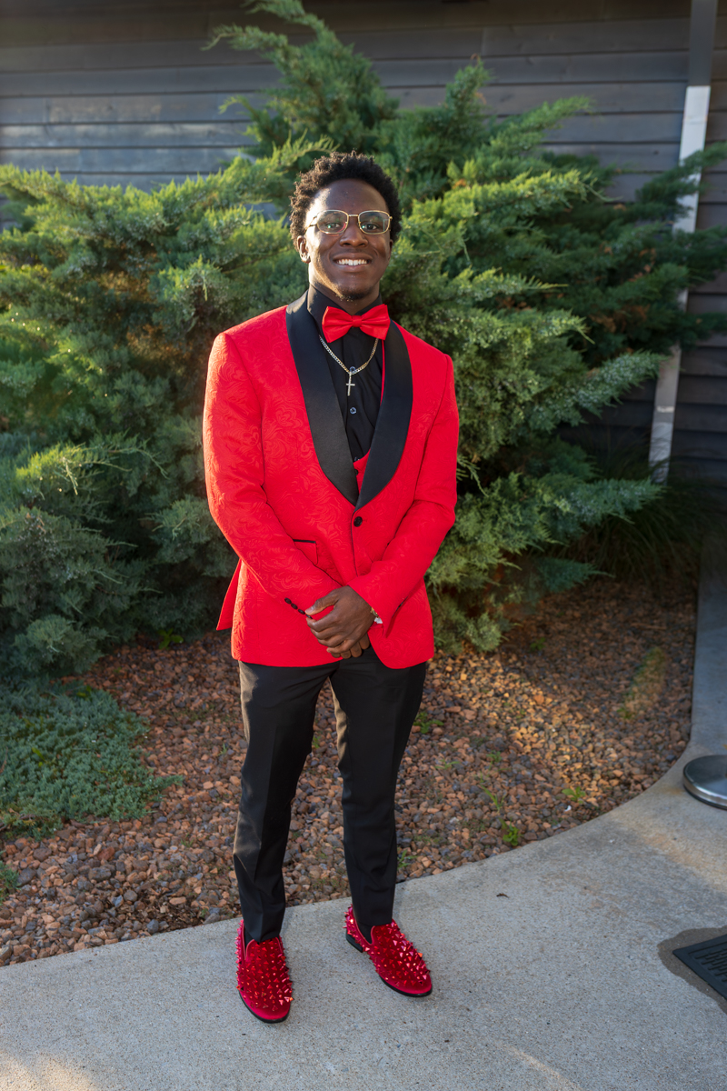 Man in a red suit jacket with black lapels, red bow tie, black pants, red studded shoes, smiling outdoors.