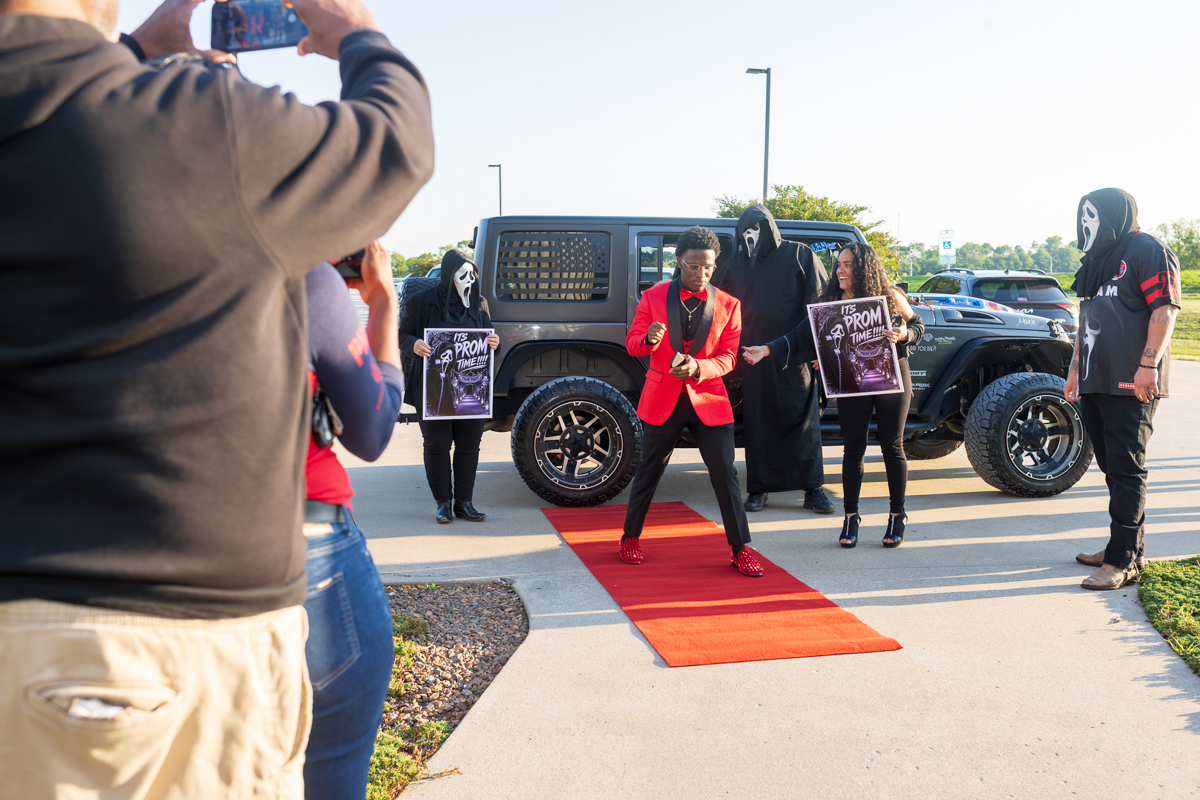 Young man in a red blazer dances on a red carpet while others in ghost masks hold prom-themed posters background a parked SUV.