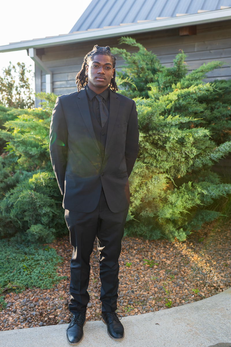 Man in a black suit posing outdoors with shrubs and a building behind it.