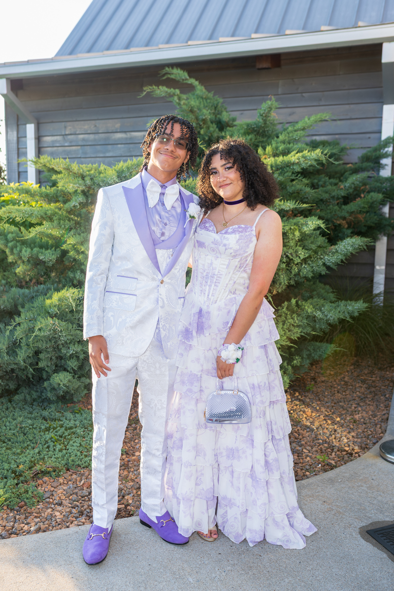 Couple in lavender formalwear posing outside a building; guy in a white suit with lavender accents and purple shoes, girl in a tiered lavender gown with a small silver purse.
