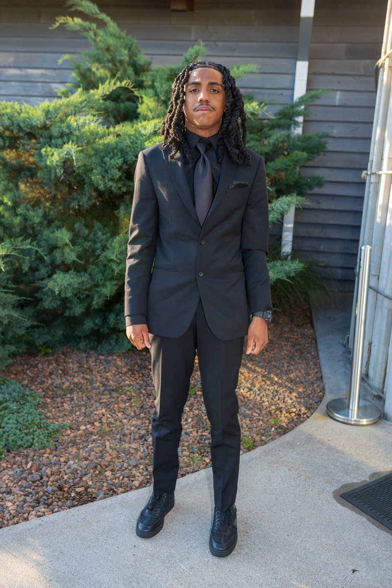 Man in a black suit and tie standing outdoors against shrubs and a wooden building backdrop.
