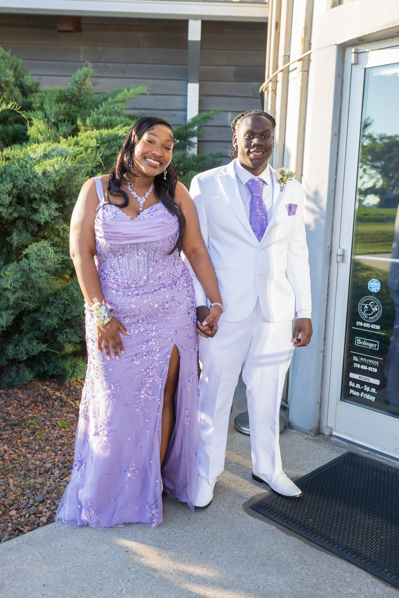 Couple dressed for a formal event, woman in a lavender gown and man in a white suit, smiling and holding hands outside a building.