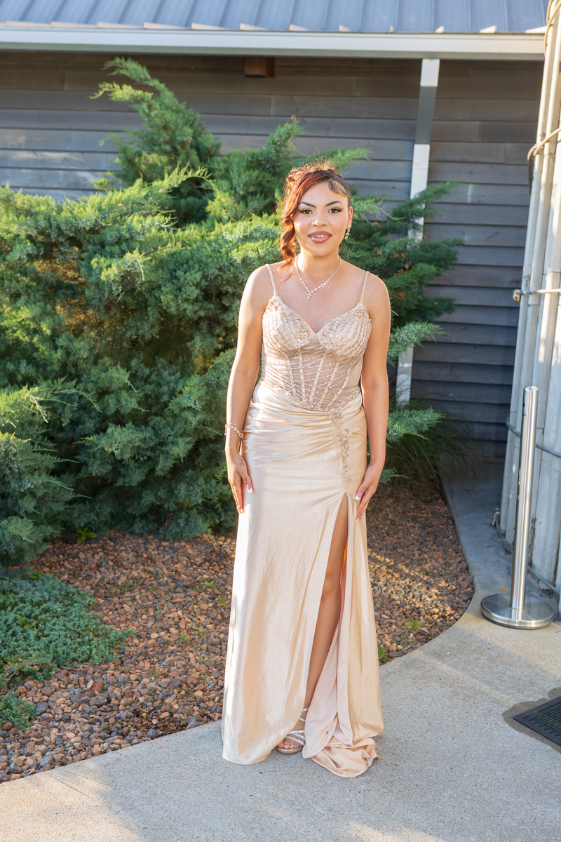 Young woman in a sparkling champagne evening gown with a thigh-high slit posing outdoors beside evergreen shrubs.