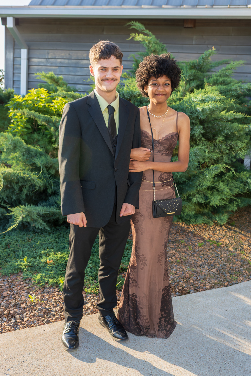 Two young adults in formal attire stand arm-in-arm outdoors, smiling by a shrubbery and house backdrop.