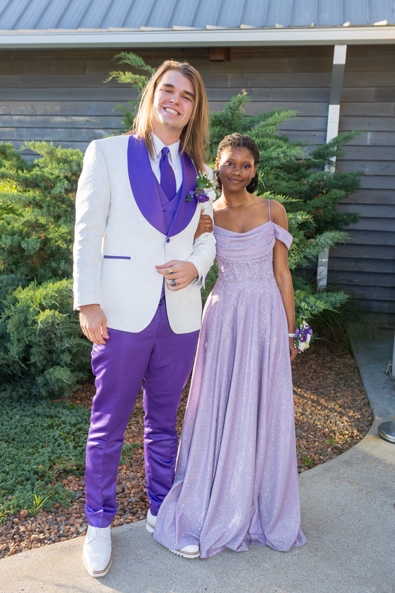 Two teenagers dressed for prom posing outdoors: the boy in a white suit with purple lapels and pants, the girl in a lavender gown, smiling together.