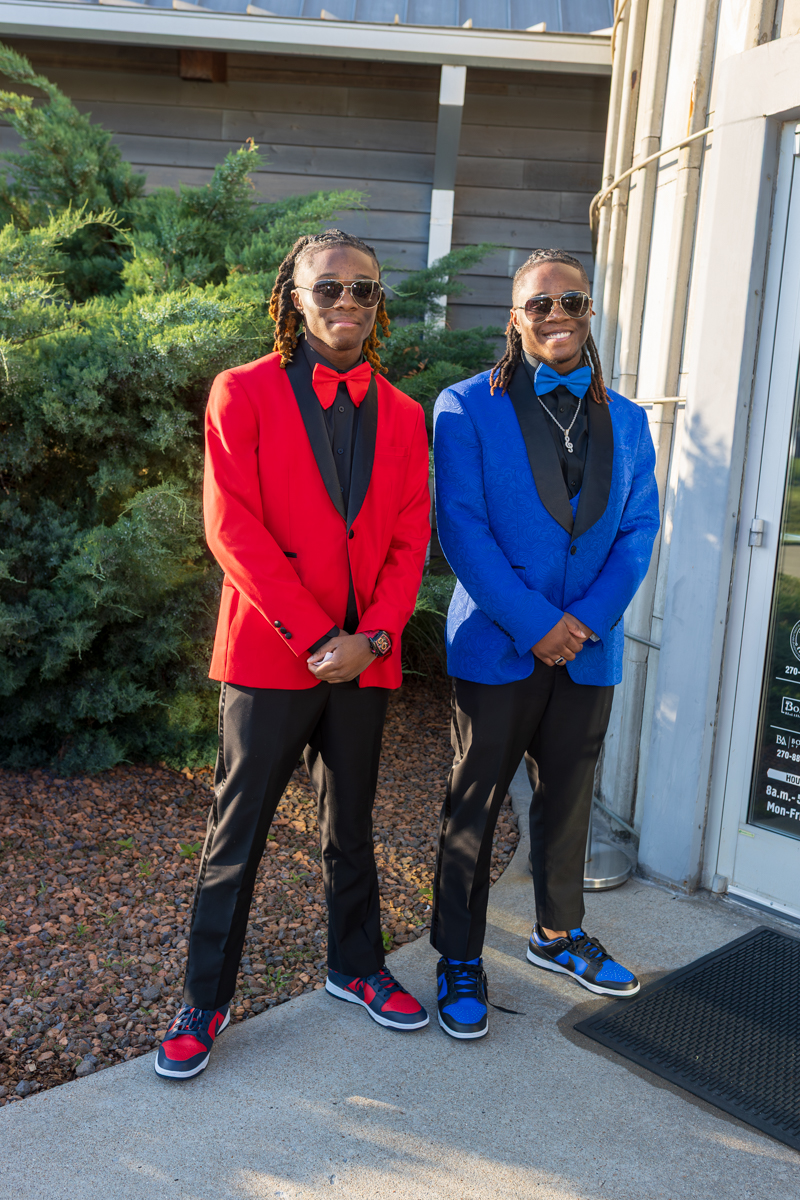 Two men in bright tuxedo jackets (red and blue) with matching bow ties and sunglasses stand outside a building with greenery in the background, smiling.
