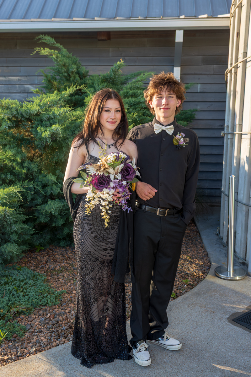 Two teens in formal wear pose outside: girl in a black beaded gown holds a purple bouquet beside a boy in a black shirt and bow tie with a boutonniere.