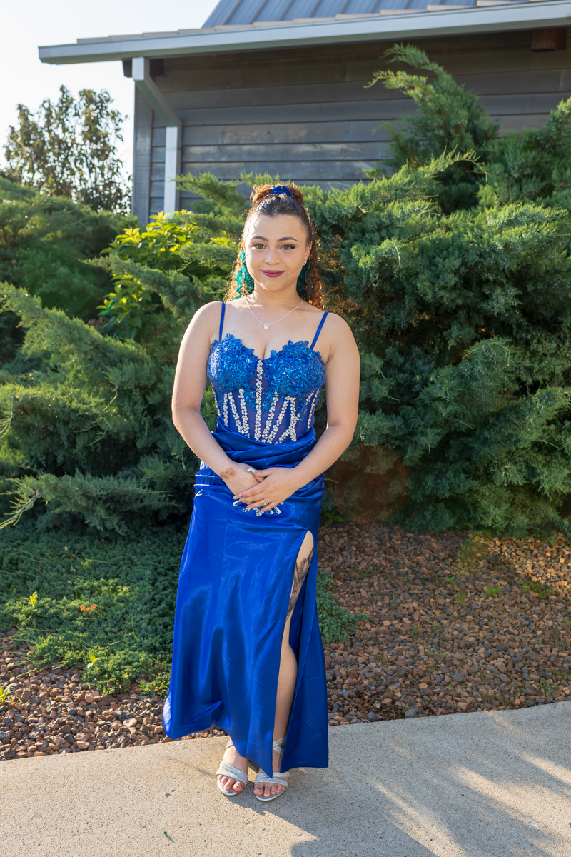 Young woman in a royal blue gown with a slit stands outdoors in front of evergreen shrubs and a building behind her to the left.