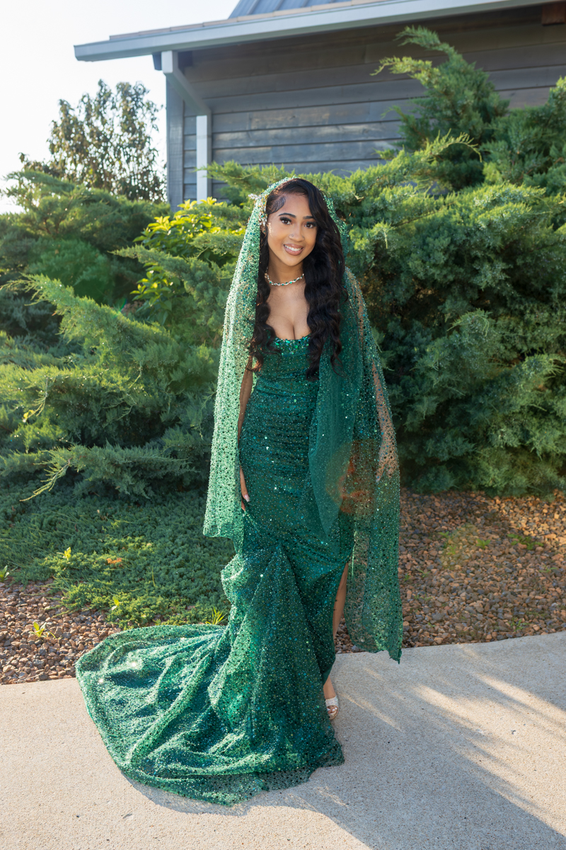 Woman in a glittering emerald green sequin gown with a matching veil standing outdoors among shrubs and a house railing behind her.