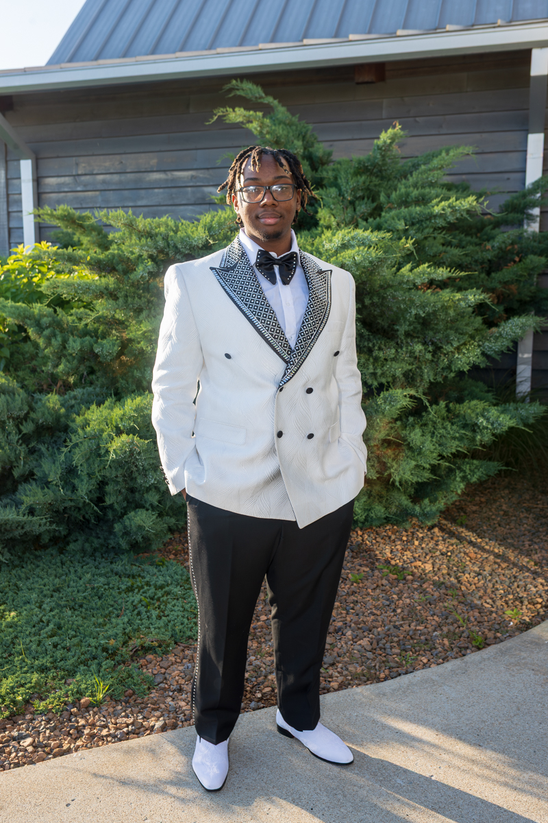 Young man wearing a white patterned tuxedo jacket with a black bow tie, black trousers, and white shoes, standing outdoors among greenery.
