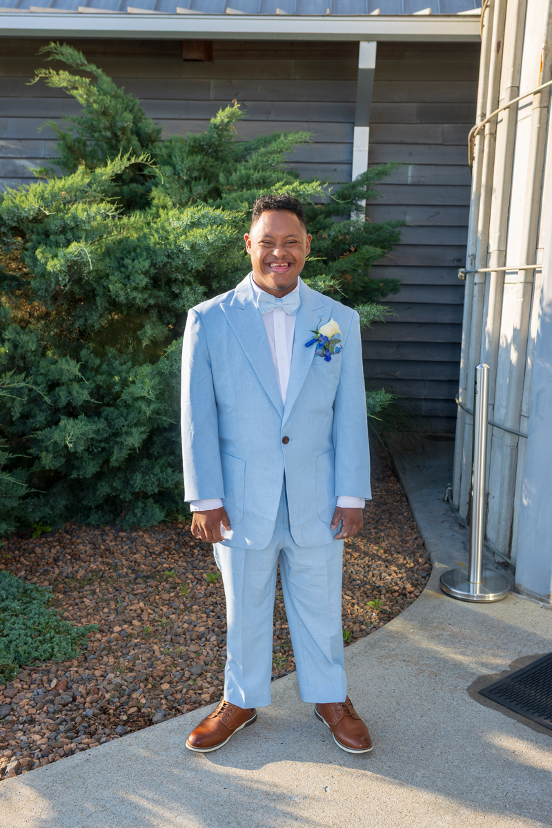 Smiling man in a light blue suit with a white bow tie and boutonniere stands on a sidewalk outdoors, greenery behind him.