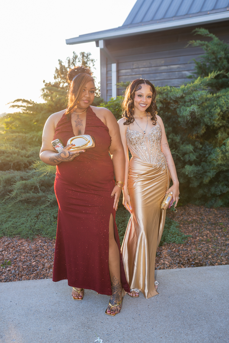 Two women in formal gowns pose together outdoors beside a house, smiling at the camera. One wears a burgundy gown with a leg slit and holds a clutch; the other wears a gold beaded dress with a matching clutch.