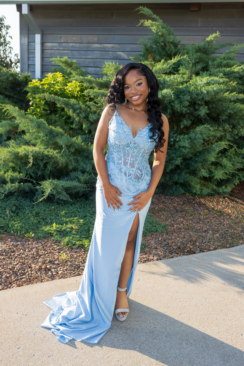 Woman in a light blue formal gown with lace bodice and high slit standing on a sidewalk outdoors, smiling at the camera.