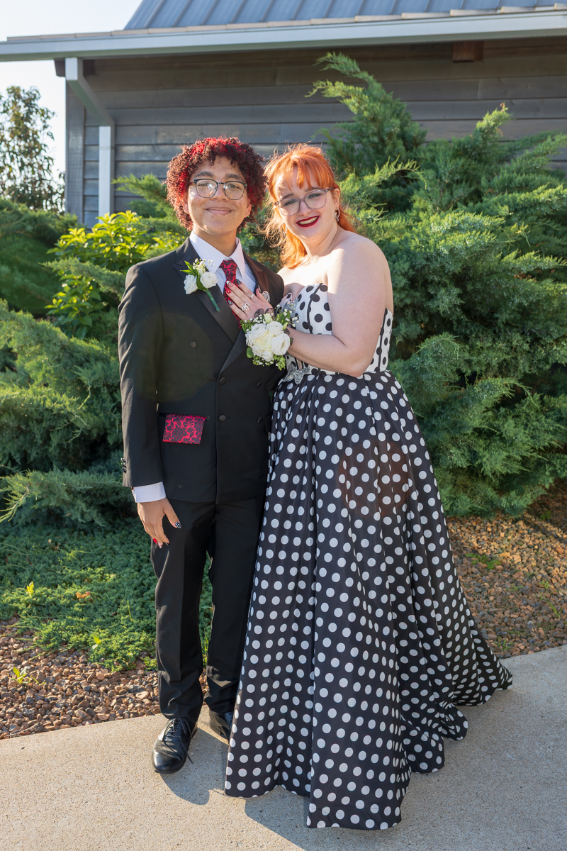 Two people in formal attire pose outdoors, smiling at the camera; a man in a black suit and a woman in a polka dot gown.