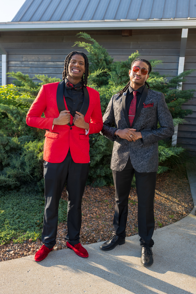 Two men in bold formal outfits pose outside a building with shrubs; one in a red blazer and red velvet shoes, the other in a dark patterned blazer with a red tie and sunglasses.