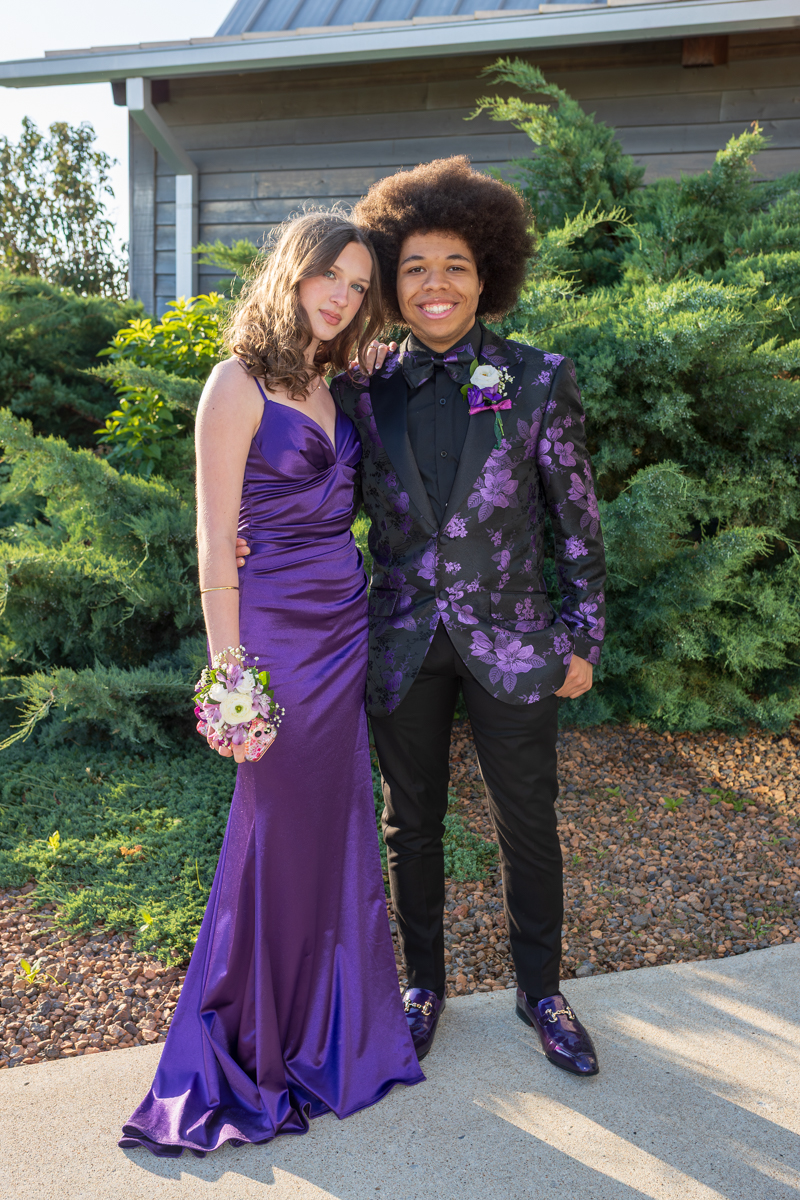 Two teens posing together outdoors in formal purple attire; the girl wears a violet gown with a corsage, the boy a black shirt with a patterned purple suit jacket and bow tie.