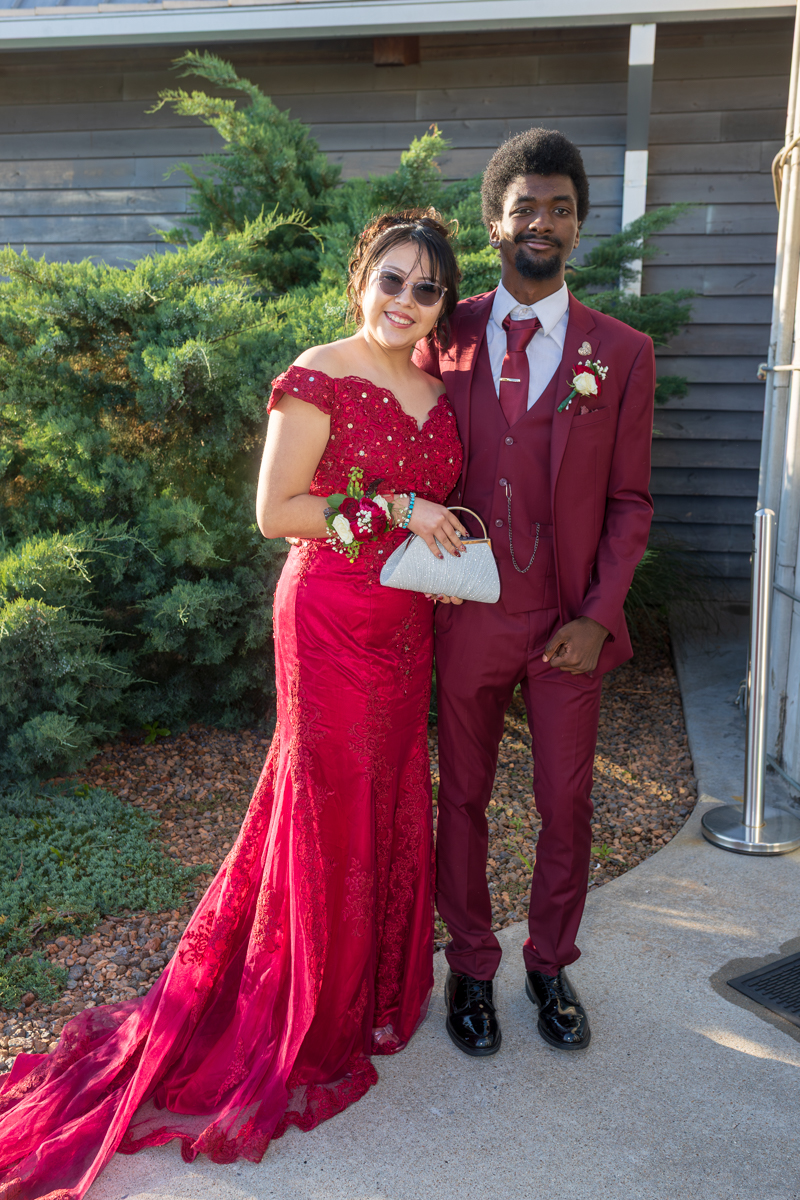 Couple dressed in formal red attire posing outdoors, the woman in a lace off-shoulder gown with a silver clutch beside a man in a burgundy suit and tie.