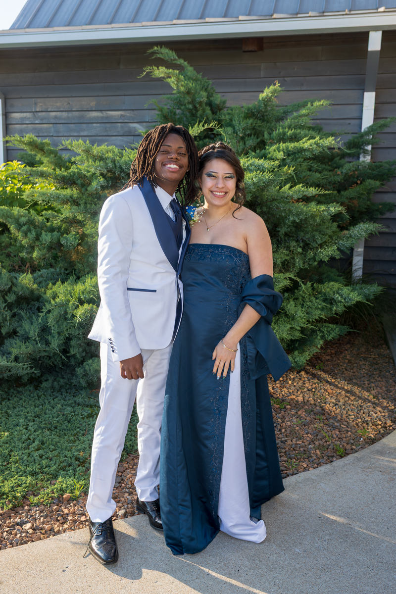 Smiling couple in formal wear posing outdoors; man in a white suit with navy accents, woman in a dark blue strapless gown with a wrap.