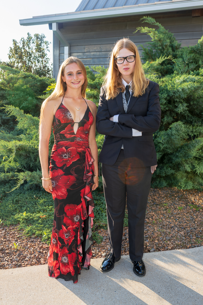 Two young women pose outdoors in formal attire: one in a red floral evening gown, the other in a dark suit with glasses.