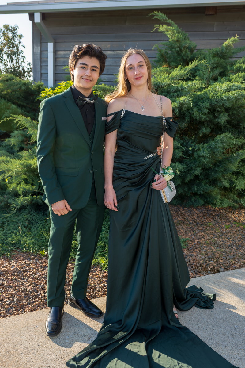 Two teens in formal attire posing outdoors: a boy in a dark green suit and a girl in a matching off‑shoulder gown with a small purse and corsage.