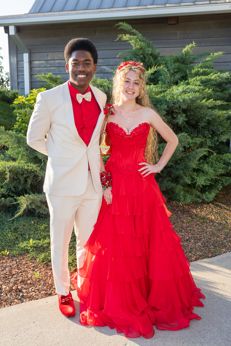 Couple in formal wear posing outdoors; man in cream suit and red shirt, woman in a red tiered gown with bouquet accessory.