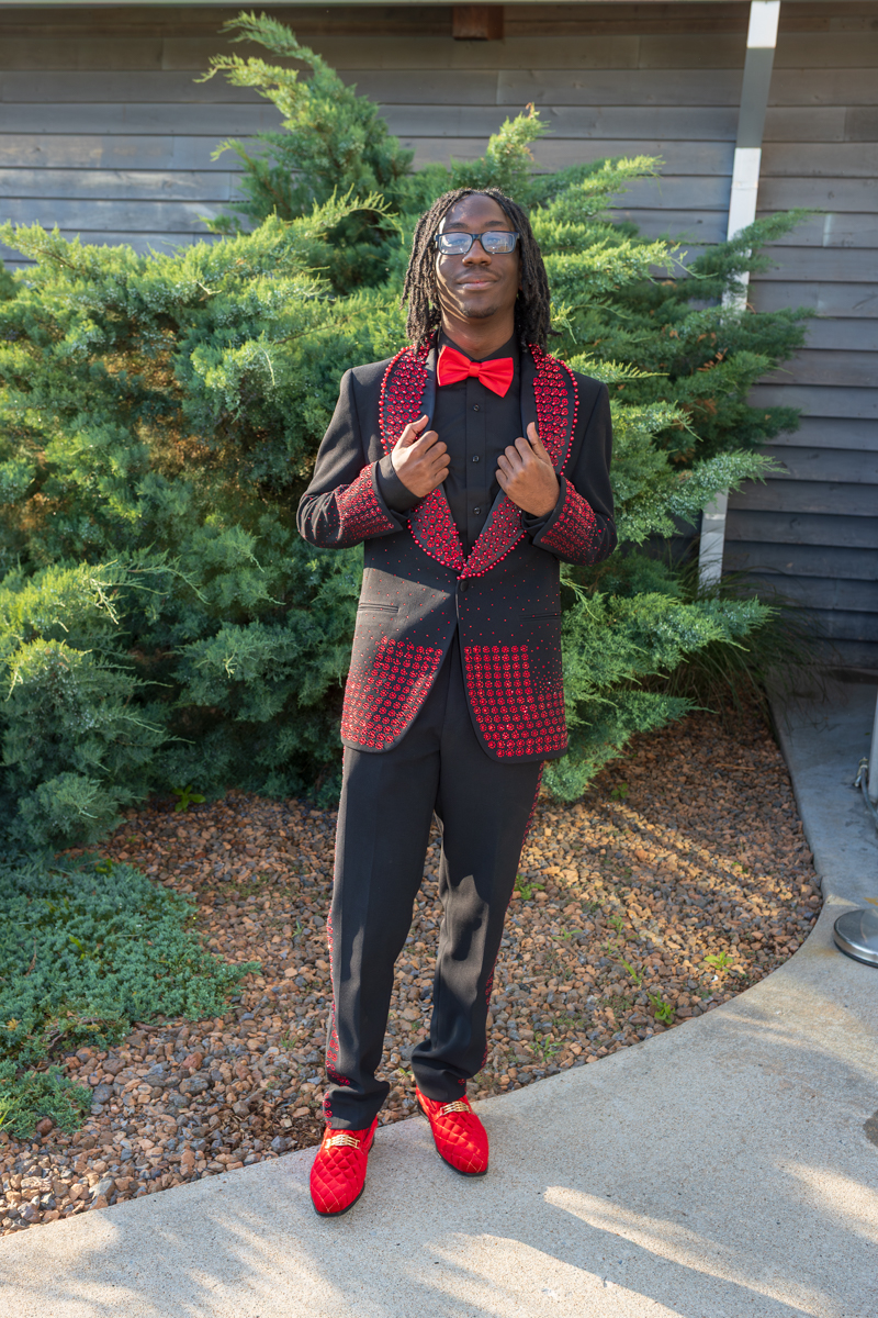 Man in a black suit with red beaded trim and red bow tie, standing outdoors in front of green shrubs, hands at chest.”