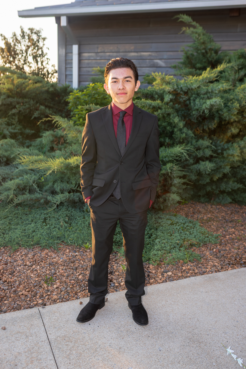 Young man in a black suit, burgundy shirt, and gray tie stands outside with bushes behind him.