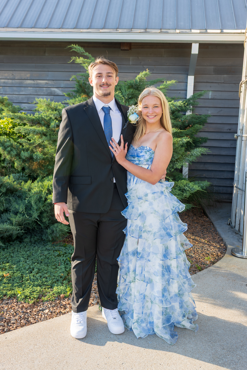 Couple in formal attire posing outdoors: man in a black suit with blue tie and white sneakers, woman in a blue floral ruffled strapless gown.