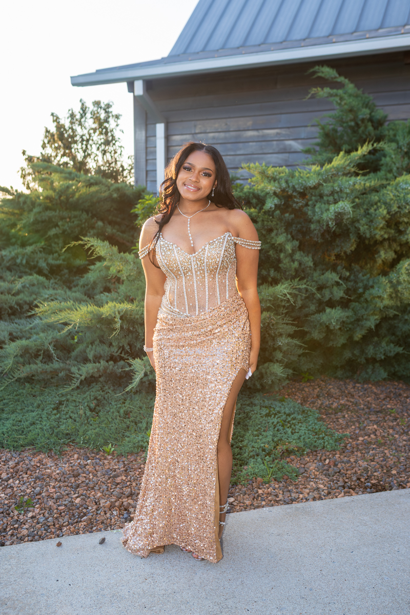 Young woman in a gold sequin evening gown with off-shoulder sleeves and a thigh-high slit, standing outdoors.