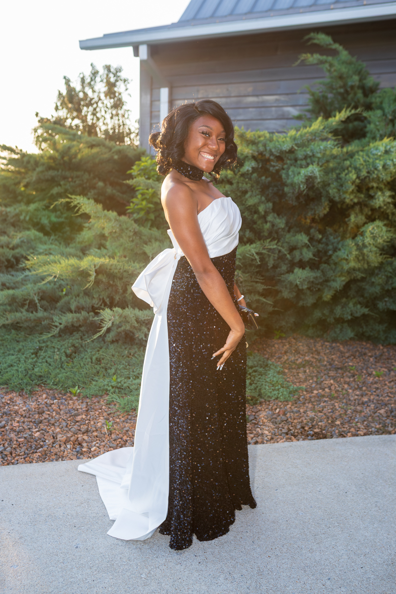 Smiling woman in a strapless black sequined gown with a white sash and train, posing outdoors.