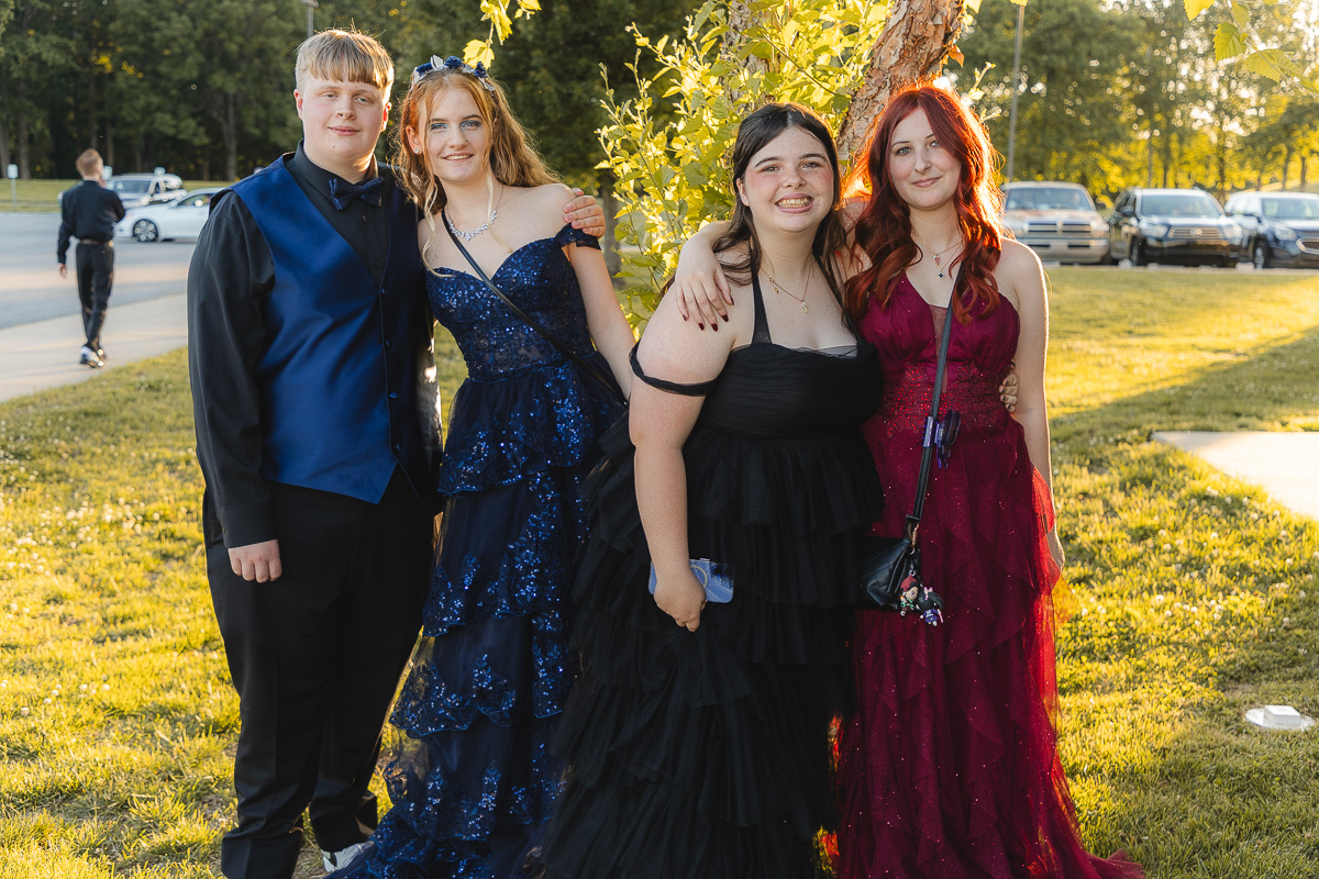 Four teens in formal attire pose together outdoors, two girls in blue gowns, one in black, one in burgundy, smiling.
