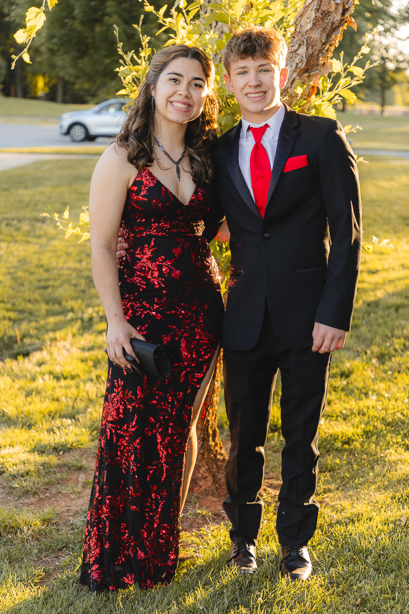 Smiling couple in formal wear outdoors by a tree: girl in a red-and-black sequined gown with a black clutch, boy in a black suit with a red tie and pocket square.
